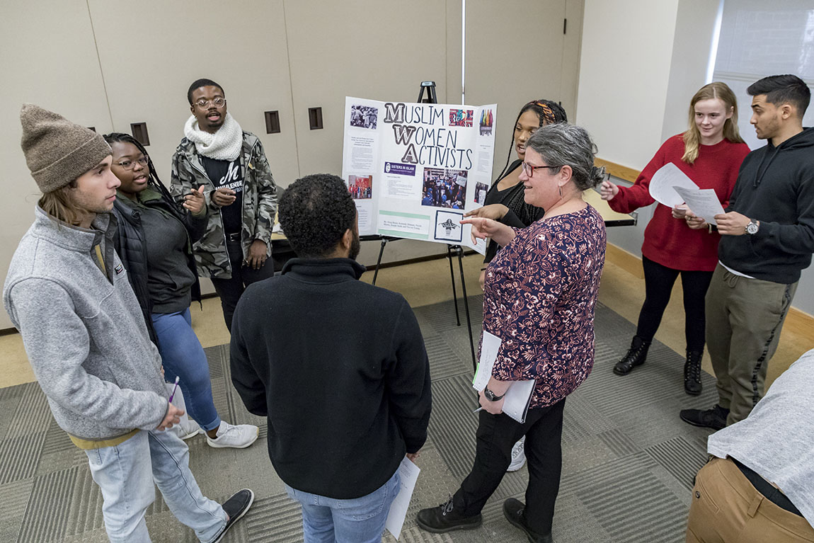 A professor speaks to a group of students, all standing around a poster board